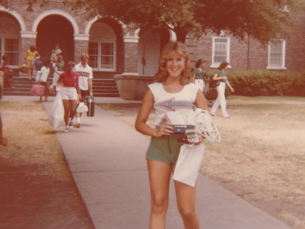Retha Stratton is a white female with blonde hair who was murdered in 1982. In this sepia toned photo, she wears green shorts and a white t-shirt and is standing on a walkway in front of a brick building on a high school campus.