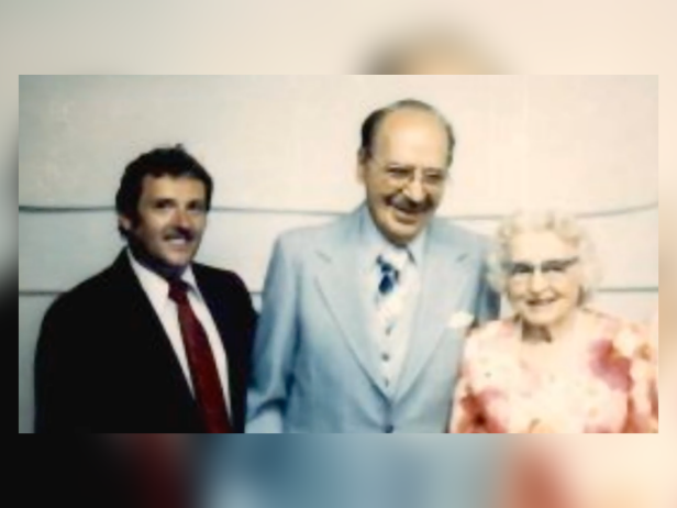 Rev. John Canning smiling next to Leo and Hazel Geese. He is in a black suit, Leo is in a blue suit, and Hazel is in a pink blouse.
