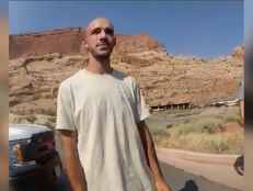 Brian Laundrie stands in the road and is wearing a white t-shirt. Behind him there are brown mountains.