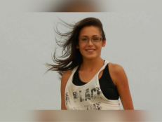 Ashley Loring/HeavyRunner stands with her hair blowing in the wind wearing black frame glasses, a white tank top and a black undershirt