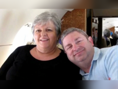 Kay and Sandy Jenkins smiling on a private plane in front of a white and brown background. Kay (left) wears a black top. Sandy (right) wears a light blue collard shirt.