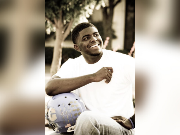 A photo of Ladarius Valentine who was murdered near his off-campus apartment in 2015. Ladarius is a Black male. In this photo he is sitting down. He is wearing a white t-shirt and jeans and is posing with a football helmet under one arm.