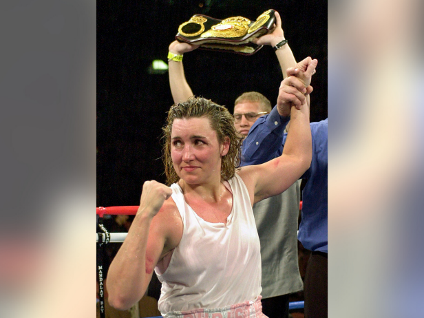Boxer Christy Martin, a white woman with blonde hair, is in a boxing ring and looks toward her corner with her right fist clenched in victory and her left arm raised over her head. Martin has on a white tank top and is drenched with sweat.