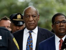 Image of Bill Cosby, an elderly Black man. He is wearing a navy suit, white shirt, and blue and brown patterned tie. He is surrounded by uniformed law enforcement officers and people in suits.