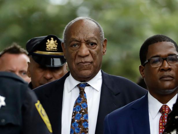 Image of Bill Cosby, an elderly Black man. He is wearing a navy suit, white shirt, and blue and brown patterned tie. He is surrounded by uniformed law enforcement officers and people in suits.