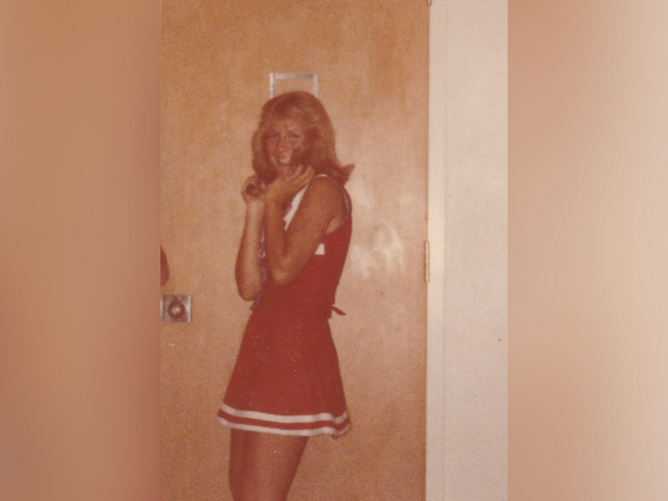 Retha Stratton is a white female with blonde hair who was murdered in 1982. In this sepia toned photo, she's wearing a red cheerleading outfit and is standing in front of a brown wood door.