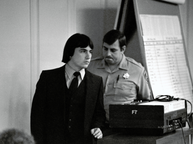 In this black and white photo, Wesley Miller, a white male with dark hair wears a dark suit and tie in a courtroom. Miller, who was convicted of the murder of Retha Stratton, is escorted by an officer of the law.