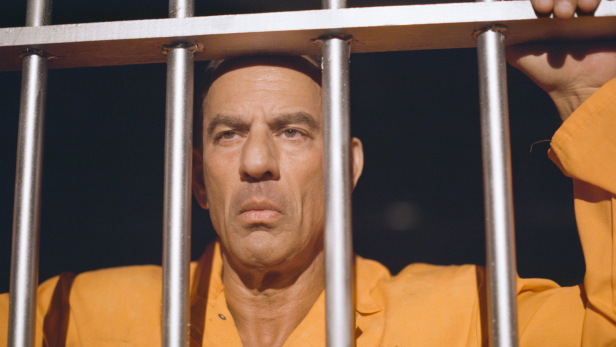 Con Man extraordinaire Barry Minkow wears an orange prison jumpsuit, stares through prison cell gates, sunlight shines through