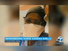 70-year-old nurse Sandra Shells smiles in an up-close selfie wearing a mask, glasses, scrubs, and a black hat. medium lighting indoors.. light and tan wall in the background.