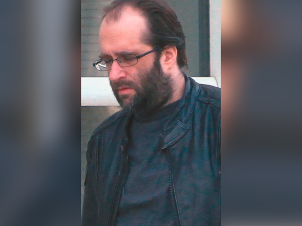 This is a photo of Jason Christopher Hughes, a white male with dark brown hair, a receding hairline, and a beard and mustache. In this image he is wearing a black t-shirt under a black jacket and black-framed eye glasses.