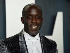 A photo of Michael K. Williams, a Black male wearing a white button down shirt under a silver and blue brocade tux jacket.