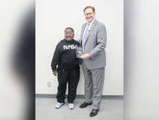 Davyon Johnson a Black male sixth grade student wears glasses, a black sweatshirt that says NASA, black sweatpants, and white sneakers. He's standing next to a white male with red hair, glasses, in a gray suit with a white shirt and light purple tie.