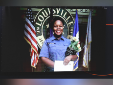 Breonna Taylor is wearing a blue button down shirt and holding a bouquet of flowers. She is standing in front of a a U.S. flag and other flags.