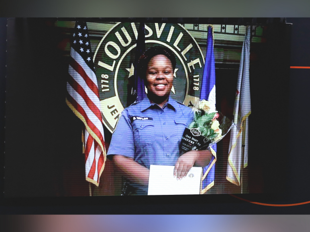 Breonna Taylor is a Black woman. In this photo she is wearing a blue button down shirt and holding a bouquet of flowers. She is standing in front of a a U.S. flag and other flags.
