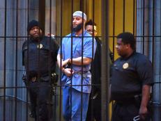 Adnan Syed is bearded, wearing a Muslim prayer cap. He is behind prison bars, handcuffed, wearing blue prison attire while being escorted by three law enforcement officers.