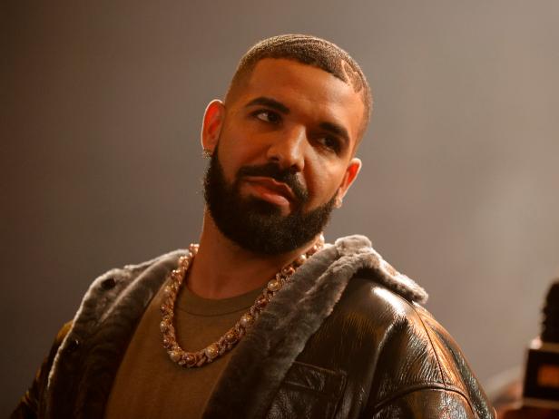 Drake is wearing a thick gold chain, a brown t-shirt, and a leather jacket with fur lining.