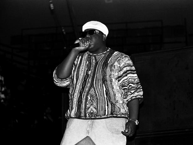 A black and white photo of Biggie Smalls aka Notorious B.I.G. He is a Black male and he is wearing a knit sweater, a white brimless hate, dark sunglasses, and light jeans. He is holding a microphone to his mouth.