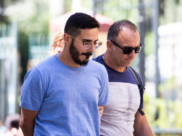 A photo of Simon Hayut aka Simon Leviev aka the "Tinder Swindler". Hayut is shown in a blue t-shirt. He is a white male with dark brown hair, a beard and mustache, wearing wire rim glasses. He is in handcuffs and escorted by a man wearing a t-shirt and dark sunglasses.