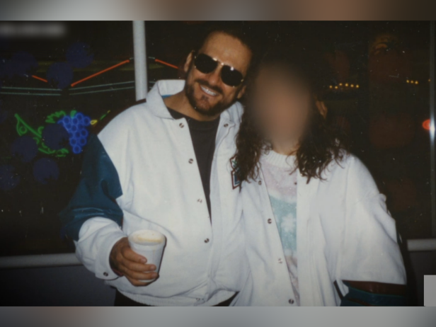 Tony Alamo–wearing a white jacket, dark sunglasses, and holding a cup–has his arm around an unidentified woman also wearing a white jacket.