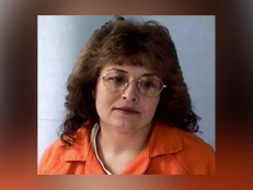 Vicki Jensen wears an orange prison top. She has brown feathered hair and large round eyeglasses.